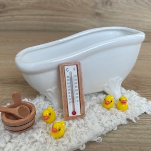 Könnte beinhalten: Eine weiße Porzellanbadewanne mit vier gelben Gummienenten, einem hölzernen Badethermometer mit einer roten Quecksilberlinie und einem Holzeimer mit einer Holzlöffel. Das Thermometer zeigt sowohl Celsius als auch Fahrenheit an.