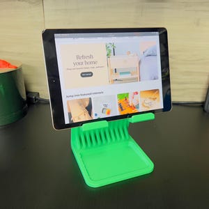 May include: A black tablet displaying a website about home decor is propped up on a bright green stand. The stand has a tray base and a comb-like support. Other items on the desk include a green container and an orange object.