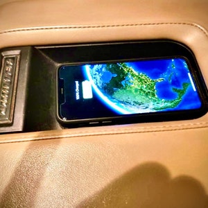 May include: A black smartphone displaying a world map is charging inside a car's center console. The console is beige leather with a black charging compartment and a silver "AMPLIFIER" emblem.