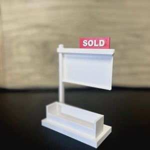 May include: A miniature white real estate sign with a red "SOLD" sign. The sign has a blank white display area and a small rectangular base. The background is blurred.