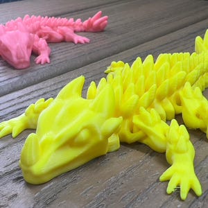 Peut inclure: Deux figurines de lézard imprimées en 3D, l'une jaune et l'autre rose, avec des détails en forme de pointes. Le lézard jaune est posé la tête tournée vers la gauche et la queue enroulée vers la droite. Le lézard rose est posé la tête tournée vers la droite et la queue enroulée vers la gauche.