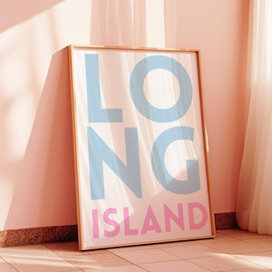 Puede incluir: Una impresión enmarcada con las palabras "LONG ISLAND" en letras azul claro y rosa sobre un fondo blanco. El marco es de madera y tiene un acabado marrón claro. La impresión se apoya en una pared rosa.