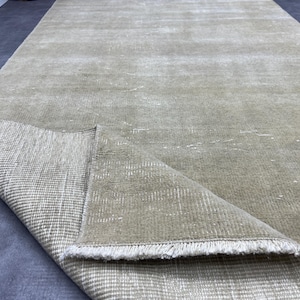 May include: A long, rectangular rug in a muted beige hue, with a distressed finish. The corner of the rug is folded over, showing a woven backing and a white fringe. The rug appears to be made of a soft, textured material, suitable for a living space.