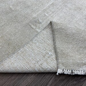 Op de afbeelding: Close-up van een beige tapijt met een opgevouwen hoek, waaruit een geweven textuur blijkt. Het tapijt heeft een franje langs één rand. De kleur is een neutraal beige met subtiele variaties, geschikt voor interieurdecoratie.
