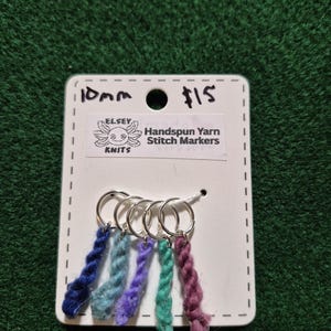Può includere: Un set di cinque segnapunti in filato filato a mano in vari colori, tra cui blu, turchese, viola e rosa. Ogni segnapunto presenta un anello argentato e un filo di filato ritorto. La scheda riporta "10mm" e "Handspun Yarn Stitch Markers."
