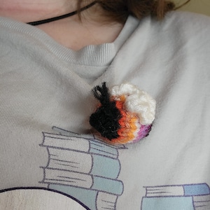 Op de afbeelding: Een handgemaakte haakspeld in de vorm van een kleine bal, met zwart, wit, oranje en paars garen. De speld zit op een lichtgrijs t-shirt met een boekenafbeelding.