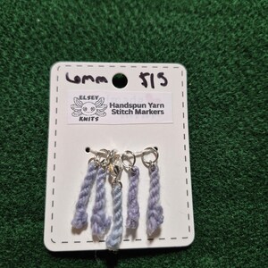 Può includere: Un set di segnapunti in filato filato a mano nei toni del blu e del viola, attaccati ad anelli color argento. I segnapunti sono esposti su una scheda bianca con il testo "6mm" e "Handspun Yarn Stitch Markers". La scheda presenta anche il logo "Elsey Knits".