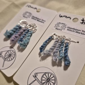 Può includere: Due set di segnapunti in filato filato a mano. Ogni set ha un'etichetta bianca con un disegno di un filatoio e il testo "Elsey Knits". I segnapunti sono realizzati in filato blu, viola e grigio e sono attaccati ad anelli d'argento.