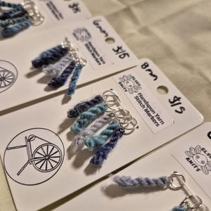 Può includere: Marcatori di punti in filato filato a mano in tonalità di blu, attaccati ad anelli d'argento. Ogni marcatore è esposto su una scheda bianca con il testo "Elsey Knits" e "Handspun Yarn Stitch Markers". Le schede hanno anche un disegno di un filatoio.