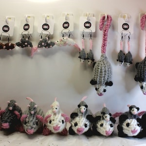 Puede incluir: Una colección de pendientes de opossum tejidos y opossums de peluche. Los pendientes están disponibles en gris, marrón y rosa. Los opossums de peluche están disponibles en gris, marrón, blanco y negro. Los opossums llevan todos una nariz rosa y tienen ojos negros.