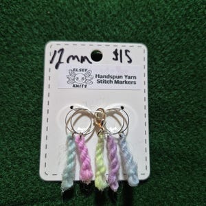 Può includere: Segnapunti in filato filato a mano su un'etichetta bianca. I segnapunti sono attaccati ad anelli argentati e presentano filato colorato nelle tonalità di blu, rosa, verde e viola. L'etichetta recita "Elsey Knits" e "12 mm".