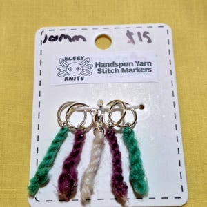 Può includere: Una scheda bianca con il testo "Handspun Yarn Stitch Markers" e il logo "Elsey Knits" mostra un set di segnapunti. Ogni segnapunto ha un anello argentato con una coda di filo colorata in verde, viola e crema.