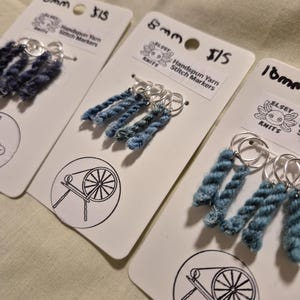 Può includere: Primo piano di tre cartoncini con segnapunti in filato filato a mano. Ogni cartoncino presenta un colore diverso di filato, fissato ad anelli argentati. I cartoncini riportano la scritta "8mm" e "16mm" e "Handspun Yarn Stitch Markers".