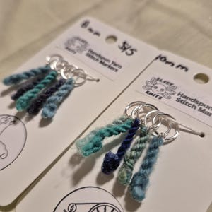 Può includere: Due set di segnapunti in filato filato a mano. Ogni set presenta un gruppo di filato blu e verde acqua attaccato a un anello d'argento. Le carte hanno il testo "Handspun Yarn Stitch Markers" e "Elsey Knits".