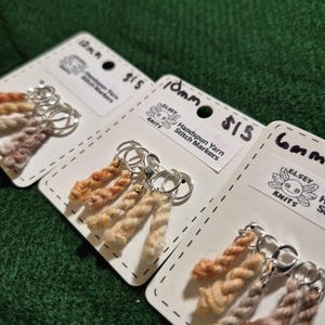 Può includere: Primo piano di tre set di segnapunti in filato filato a mano su cartoncini bianchi. Ogni set ha un colore di filato diverso ed è attaccato a un anello d'argento. Le carte hanno il testo "ELSEY KNITS Handspun Yarn Stitch Markers".