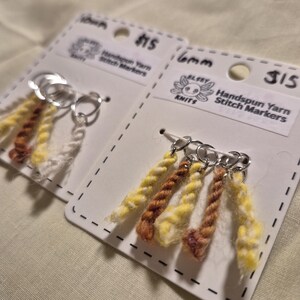 Può includere: Due set di segnapunti in filato filato a mano su cartoncini bianchi. Ogni segnapunti presenta un anello argentato con tre fili di filato giallo, marrone e bianco. Le carte hanno il testo "Handspun Yarn Stitch Markers" e "ELSEY KNITS".