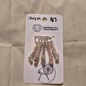 Può includere: Segnapunti in filato filato a mano su una scheda bianca. La scheda ha il testo "10mm" e "$15" scritto sopra. I segnapunti sono attaccati ad anelli d'argento. Il filato è un mix di colori rosa, marrone e blu. La scheda ha un logo di un filatoio.