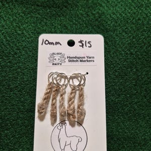 Può includere: Un'etichetta bianca con la scritta "10mm" e "$15" mostra dei segnapunti in filato filato a mano. I segnapunti sono attaccati ad anelli argentati e hanno un aspetto intrecciato marrone chiaro. L'etichetta presenta anche un'illustrazione di un lama.