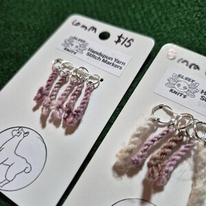 Può includere: Due cartoncini bianchi con segnapunti in filato filato a mano. Ogni cartoncino ha cinque segnapunti in filato nelle tonalità del rosa e del crema, attaccati ad anelli argentati. I cartoncini presentano un logo con un lama e il testo "Handspun Yarn Stitch Markers".