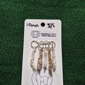 Può includere: Una scheda bianca con il testo "10mm 5/5" e "Handspun Yarn Stitch Markers" con un logo di lama. Sono attaccati quattro segnapunti con filato crema e marrone. Lo sfondo è verde.