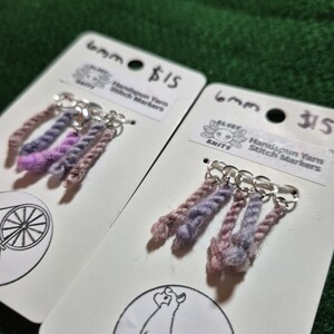 Può includere: Due segnapunti in filato filato a mano su cartoncini bianchi. Ogni segnapunto presenta un gruppo di fili colorati nei toni del viola, rosa e marrone. I cartoncini mostrano il testo "6mm" e "Handspun Yarn Stitch Markers".