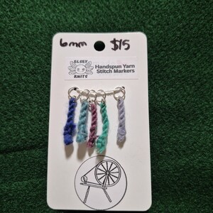 Può includere: Un cartoncino bianco con sei segnapunti in filato filato a mano nei toni del blu, verde e viola. La scheda ha il testo "6mm" e "Handspun Yarn Stitch Markers". Un logo di un filatoio è anche sulla scheda.