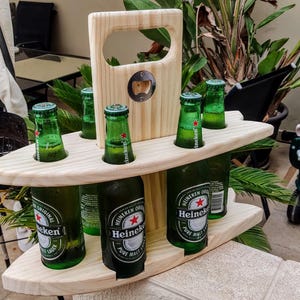 May include: Wooden six-pack beer holder with a built-in bottle opener. The holder is shaped like a surfboard and has a handle. It is filled with six green glass bottles of beer, including Heineken.