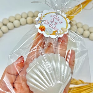 May include: A clear cellophane bag tied with a silver ribbon, containing a white seashell-shaped item. A tag with the text "Ada's First Birthday" and seashell graphics is attached. The background includes decorative beads and dried wheat.