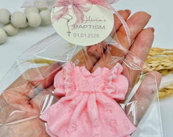May include: A pink dress-shaped soap favor for a baptism, packaged in a clear cellophane bag and tied with a pink ribbon. The tag reads "Olivia's BAPTISM 01.01.2026". The soap has a detailed floral design.