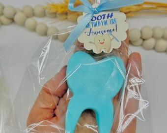 May include: A light blue tooth-shaped item in a clear cellophane bag, tied with a light blue ribbon. A tag with the text "Tooth Be Told You Are Awesome!" is attached. The item is held in a hand.