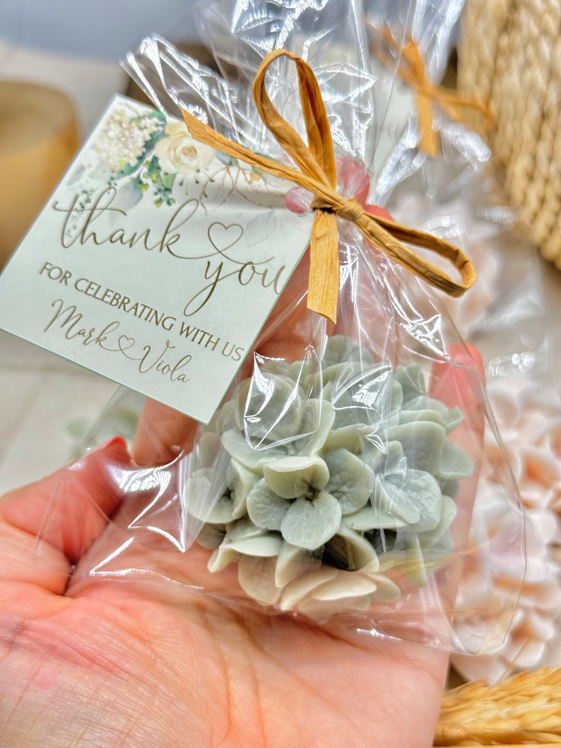 May include: A small, white and green floral arrangement in a clear plastic bag tied with a brown ribbon. A thank you tag with the text "Thank you for celebrating with us Mark & Viola" is attached to the bag.