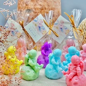 May include: A collection of colorful astronaut-shaped soaps, each seated on a textured moon. The soaps are in various pastel colors, including yellow, pink, green, blue, and purple. Some are packaged in clear cellophane bags with tags that read "Thank you for making my party a blast."