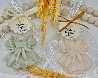 May include: Two baby dress-shaped soaps in clear cellophane bags, tied with ribbons and tags that read "Phoebe's Baptism 01.10.23." One soap is pale green, the other is light pink. Wheat stalks and wooden beads are in the background.