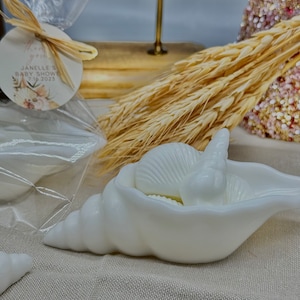 May include: White seashell shaped soap dish with a white seashell soap inside. The soap dish is on a white fabric surface.