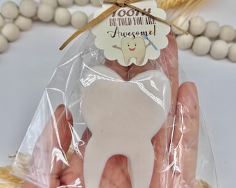 May include: A white tooth-shaped item is packaged in a clear cellophane bag, tied with a raffia bow. A small tag reads "TOOTH BE TOLD YOU ARE Awesome!" with a cartoon tooth illustration. The item is held in a hand.