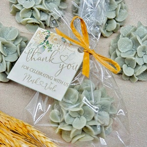 May include: A set of five small, light green hydrangea flower-shaped favors in clear cellophane bags tied with a yellow ribbon. A small card with the text "thank you" and "for celebrating with us Mark & Viola" is attached to the ribbon.