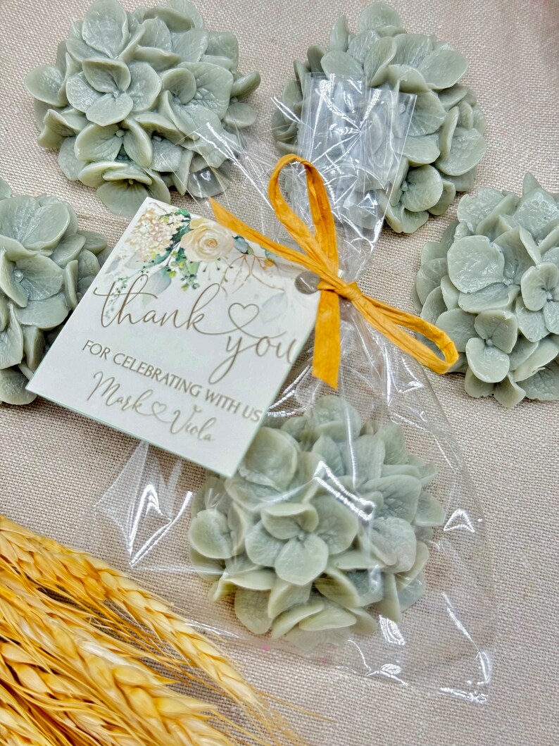 May include: A set of five small, light green hydrangea flower-shaped favors in clear cellophane bags tied with a yellow ribbon. A small card with the text "thank you" and "for celebrating with us Mark & Viola" is attached to the ribbon.