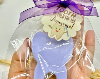 May include: A light purple tooth-shaped item is wrapped in clear cellophane and tied with a purple ribbon. A small tag with the text "TOOTH BE TOLD YOU ARE Awesome!" and a cartoon tooth graphic is attached. The item is a party favor.