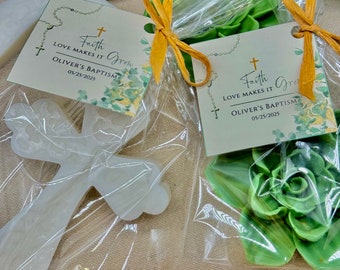 May include: Two white cross-shaped favors wrapped in clear cellophane bags with a gold ribbon. Each bag contains a green succulent and a card with the text "Faith Grow Love Makes It Oliver's Baptism 05/25/2025".