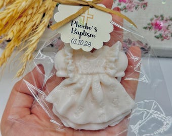 May include: A small, white dress-shaped soap is packaged in a clear cellophane bag, tied with a raffia bow. A tag reads "Phoebe's Baptism 01.10.23". The soap has a ruffled neckline and a detailed floral pattern.