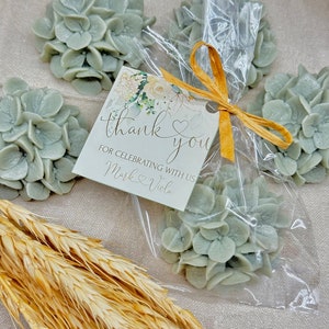 May include: A small clear bag filled with four light blue hydrangea flower arrangements. A white card with the text "thank you" and "for celebrating with us Mark & Viola" is tied to the bag with a gold ribbon.
