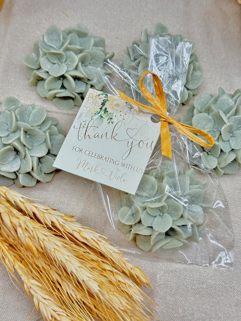 May include: A small clear bag filled with four light blue hydrangea flower arrangements. A white card with the text "thank you" and "for celebrating with us Mark & Viola" is tied to the bag with a gold ribbon.