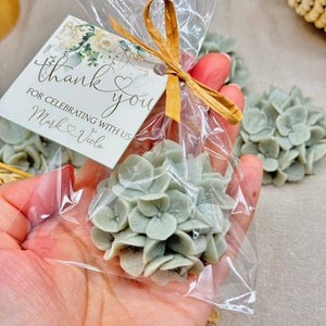May include: A small clear bag filled with a light green hydrangea flower. The bag is tied with a brown ribbon and has a thank you tag attached that reads "thank you for celebrating with us Mark & Viola".