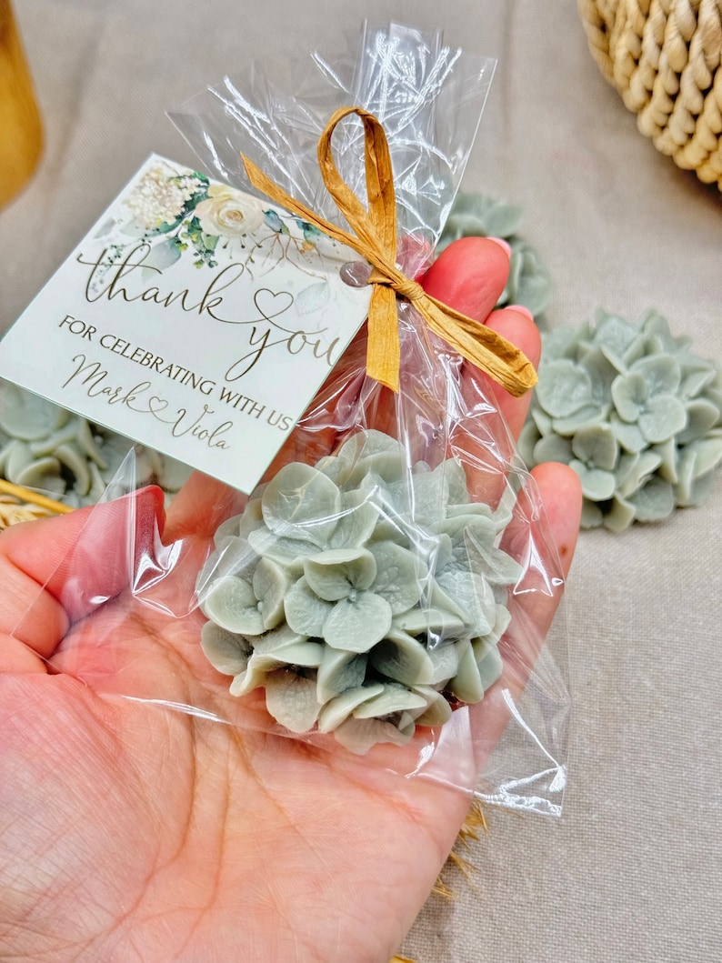 May include: A small clear bag filled with a light green hydrangea flower. The bag is tied with a brown ribbon and has a thank you tag attached that reads "thank you for celebrating with us Mark & Viola".