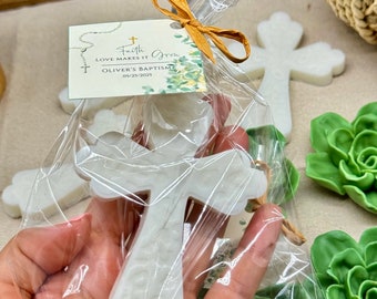 May include: White cross-shaped soap favors in a clear plastic bag with a gold ribbon and a tag that reads "Faith Love Makes It Grow Oliver's Baptism 05/23/2023".