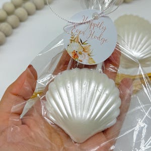 May include: A pearlescent white seashell-shaped soap is packaged in a clear cellophane bag. A round tag with the words "Happily Ever Hodge" and floral accents is attached. The soap is held in a person's hand.