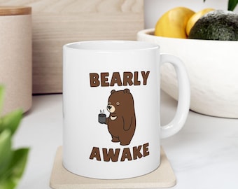 Op de afbeelding: Witte keramische mok met de woorden "BEARLY AWAKE" in bruine letters en een cartoonbeer met een koffiekopje. De mok staat op een beige onderzetter.
