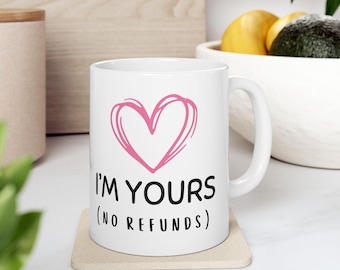Può includere: Tazza in ceramica bianca con un cuore rosa e la scritta "I'M YOURS (NO REFUNDS)". La tazza è appoggiata su un sottobicchiere beige.