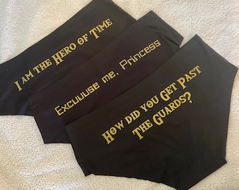 May include: Black fabric with gold text. The phrases "I AM THE HERO OF TIME", "Excuuuse me, Princess", and "HOW DID YOU GET PAST THE GUARDS?" are printed on the fabric. The fabric is folded and arranged on a light-colored surface.