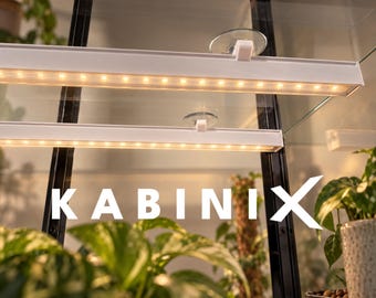 Può includere: Due barre luminose a LED bianche montate con ventose trasparenti all'interno di una vetrinetta in vetro. Le luci emettono un bagliore caldo sulle piante verdi. Il testo KABINIX è visualizzato in lettere maiuscole bianche al centro.